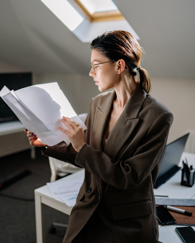 Woman in a brown check blazer reads scattered papers at a desk near a skylight in a bright office.